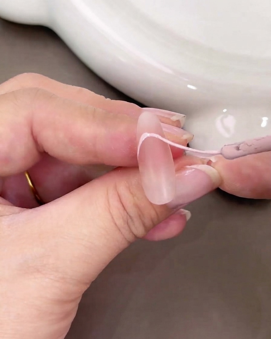 Gros plan dune main utilisant un pinceau fin pour peindre une courbe de French tip rose clair sur un ongle nude.