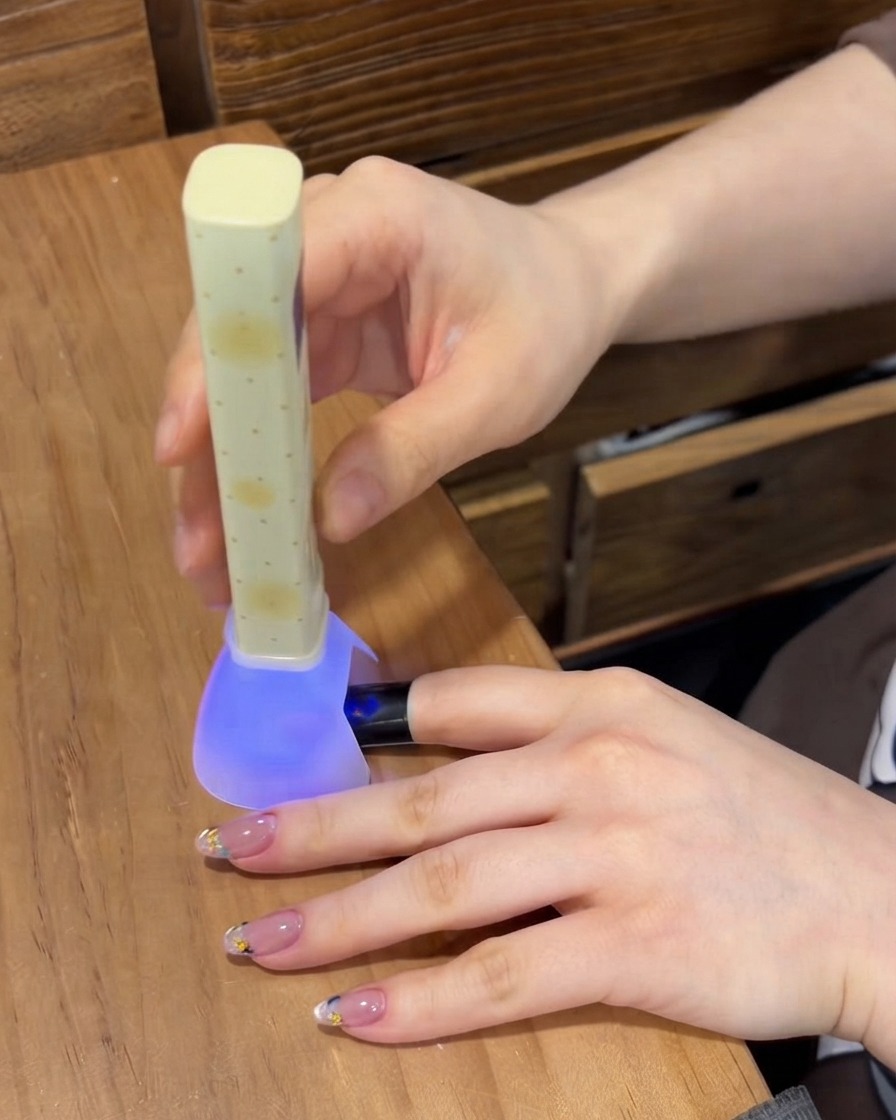 Une mini lampe UV/LED violette portable catalysant le gel sur un ongle tandis que dautres doigts manucurés reposent sur la table.