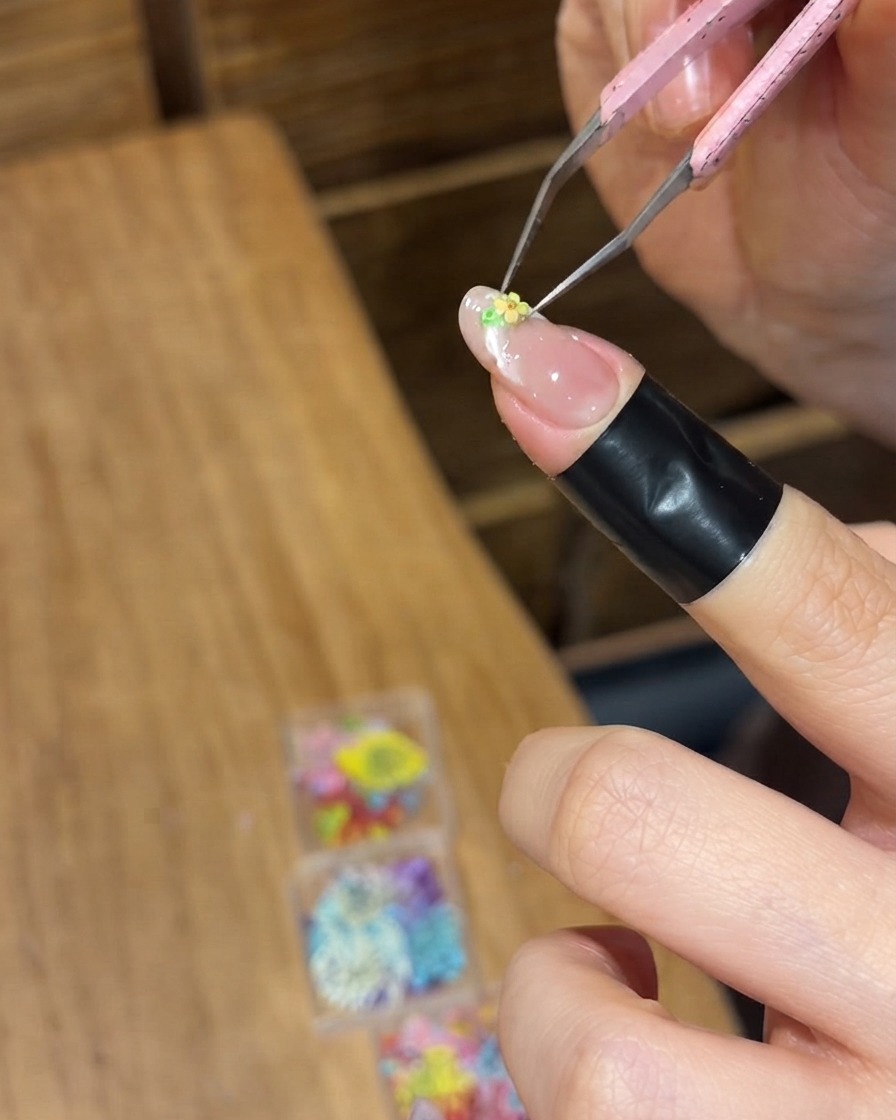 Une pince de précision positionnant une minuscule fleur séchée jaune sur la surface en gel humide dun ongle.