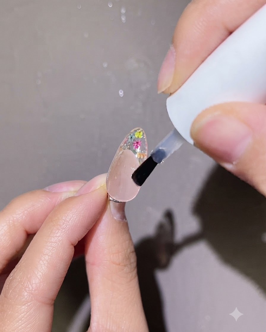 Un petit pinceau appliquant une goutte précise de top coat transparent sur le nail art floral dun onglet.