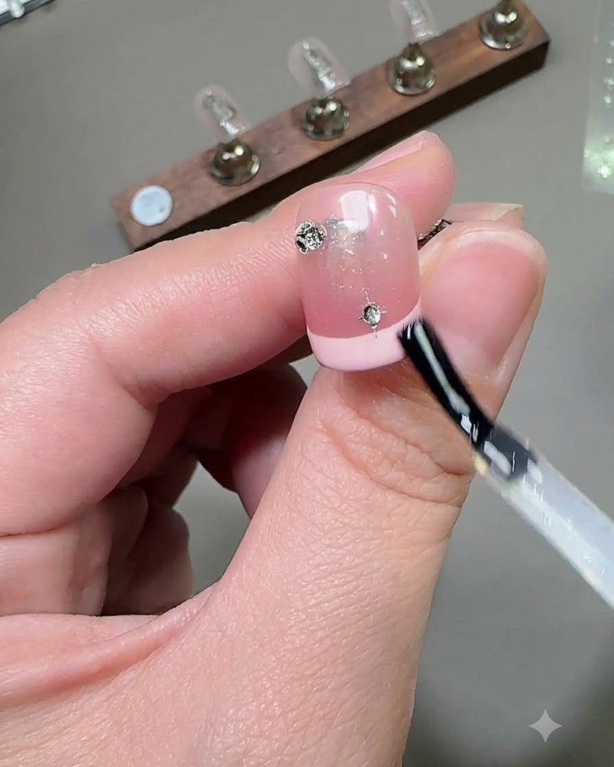 Un pinceau appliquant une couche de finition brillante sur un ongle décoré de strass et de charms étoiles.