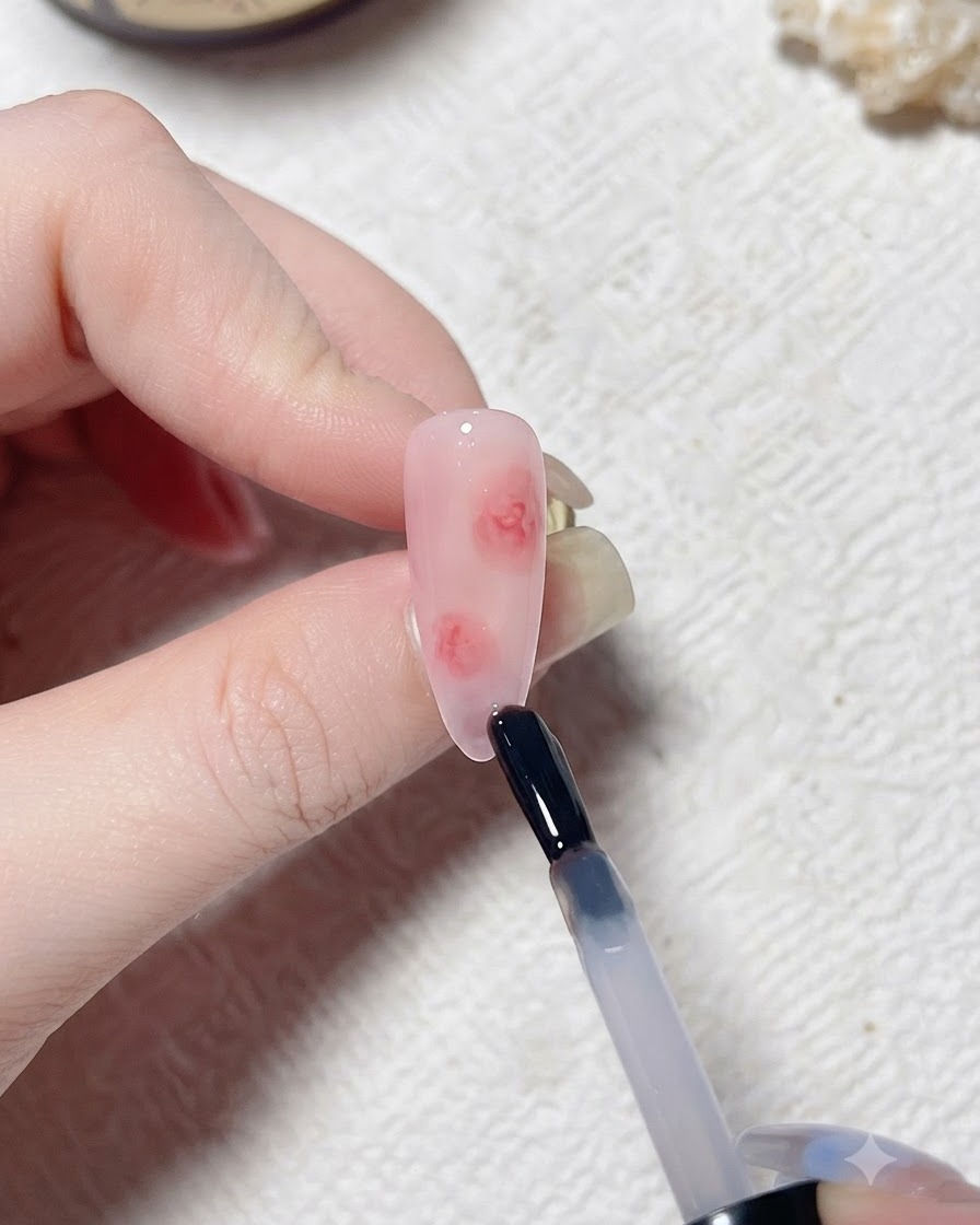 Un pinceau appliquant une couche transparente de blooming gel sur un faux ongles aux tons roses.