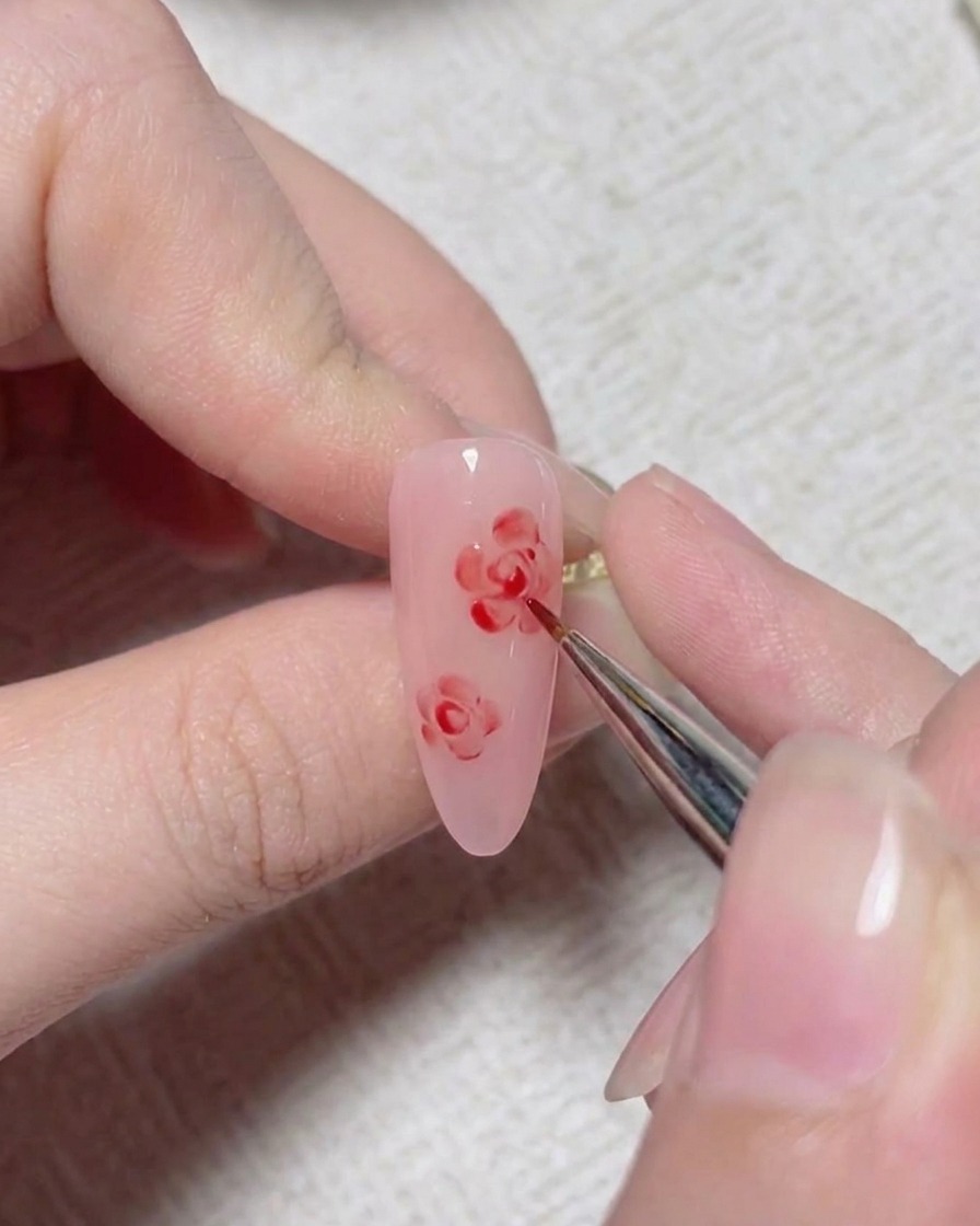 Un pinceau fin de nail art ajoutant des lignes de détail rouges pour former le centre dune rose peinte sur un ongle rose.