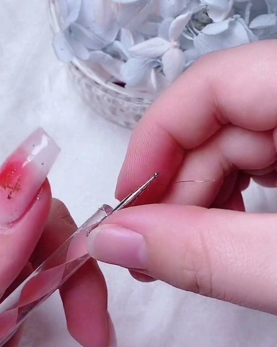 Une pince fine plaçant une petite rose complexe en fil doré au centre dun ongle décoré.
