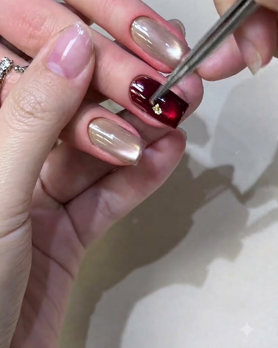 Une paire de pinces plaçant de petits morceaux de feuille dor sur un ongle verni au gel bordeaux.