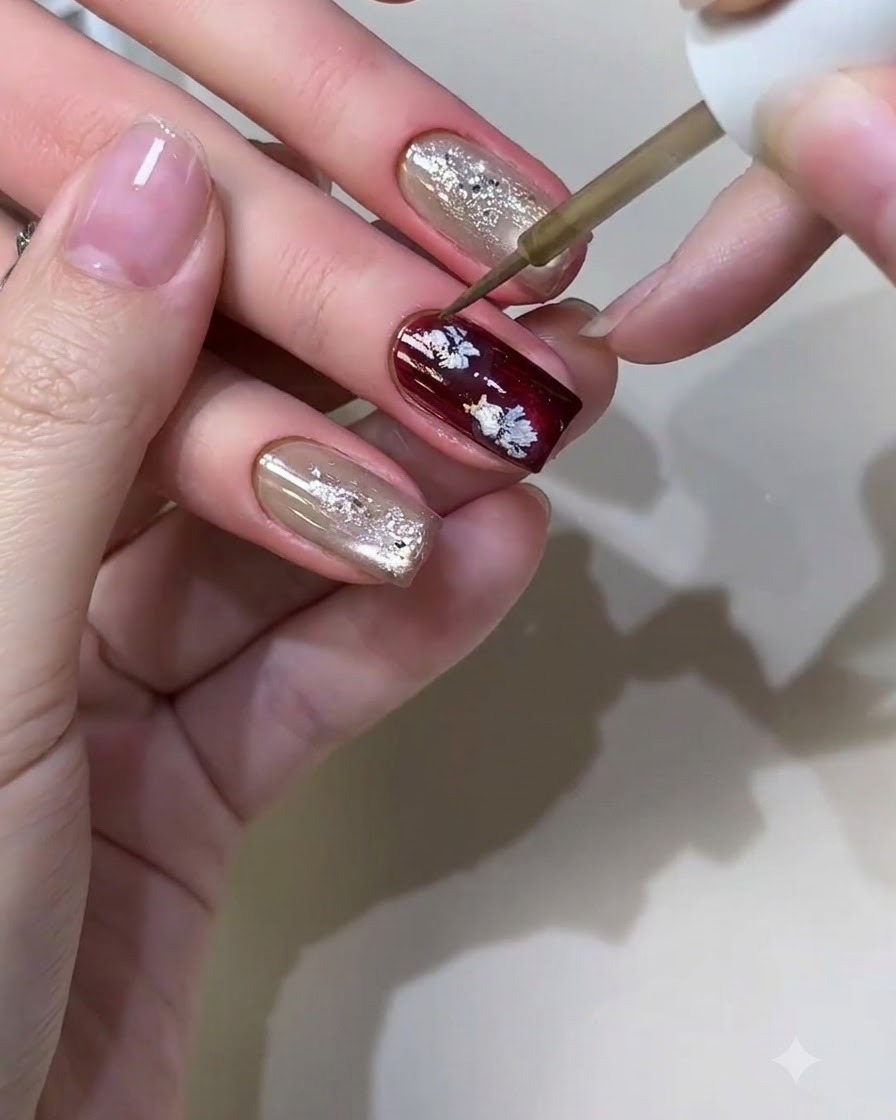 Un pinceau à nail art fin appliquant du liquide métallique pour former de petits motifs floraux décontractés sur un ongle bordeaux.