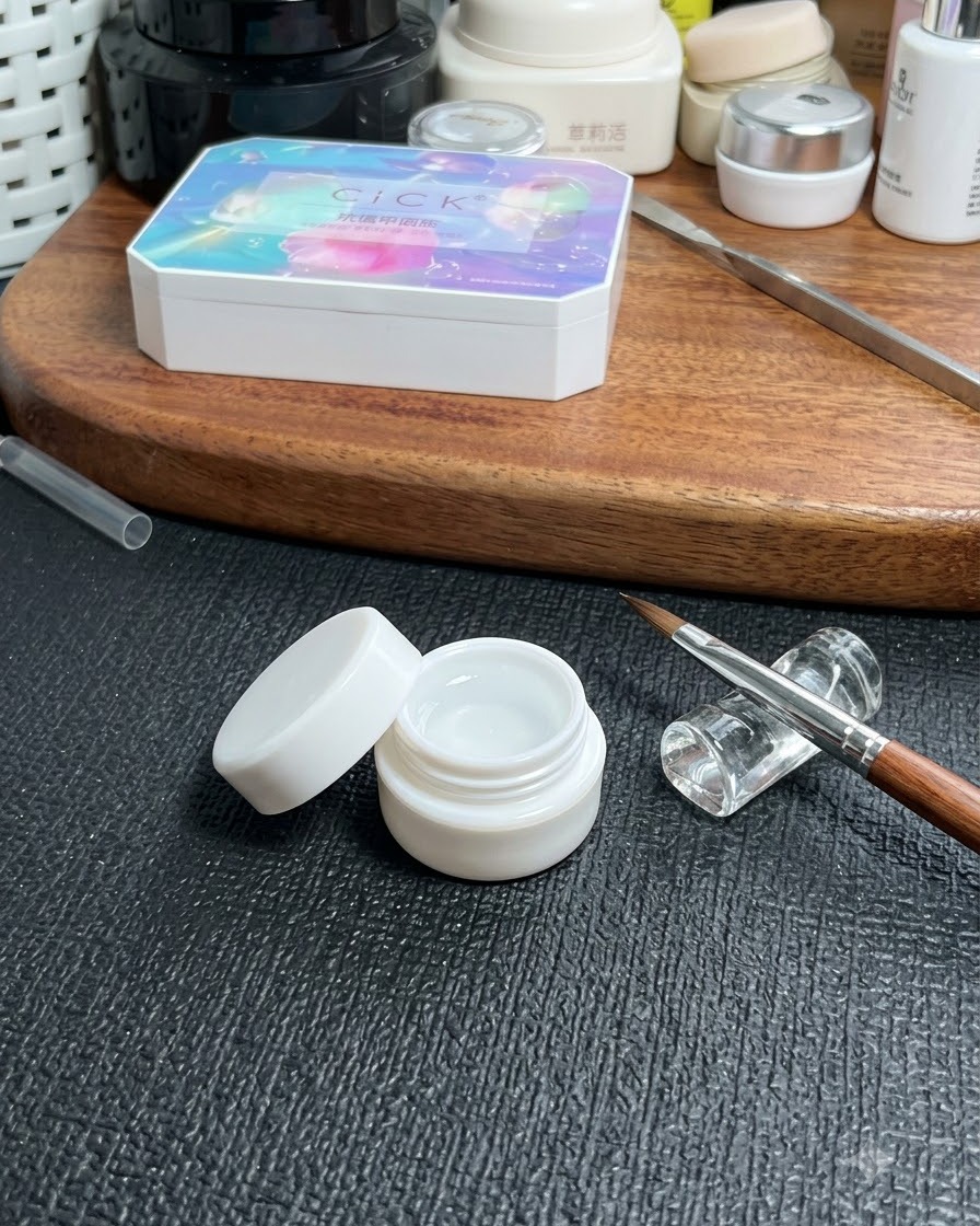 Un pot de gel de base transparent placé sur une table à côté dun pinceau fin pour nail art et dune boîte de rangement cosmétique.