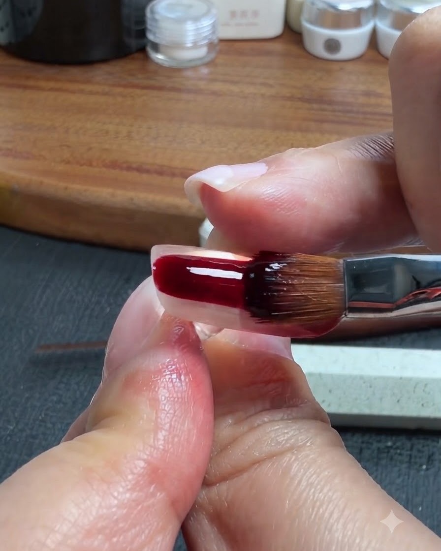 Un pinceau à ongle appliquant un gel translucide rouge-brun foncé sur la partie inférieure dun ongle artificiel transparent.