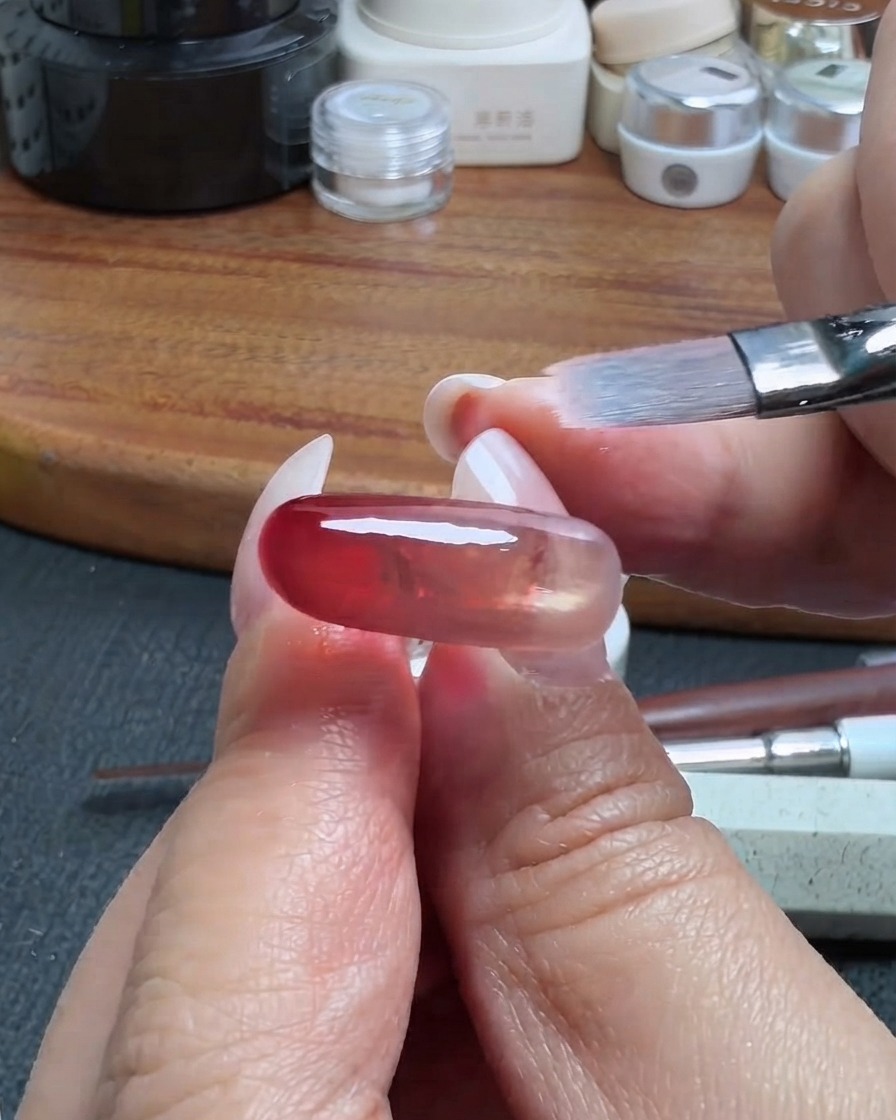 Un pinceau de maquillage propre estompant délicatement le bord dune couche de gel translucide rouge sur un ongle.