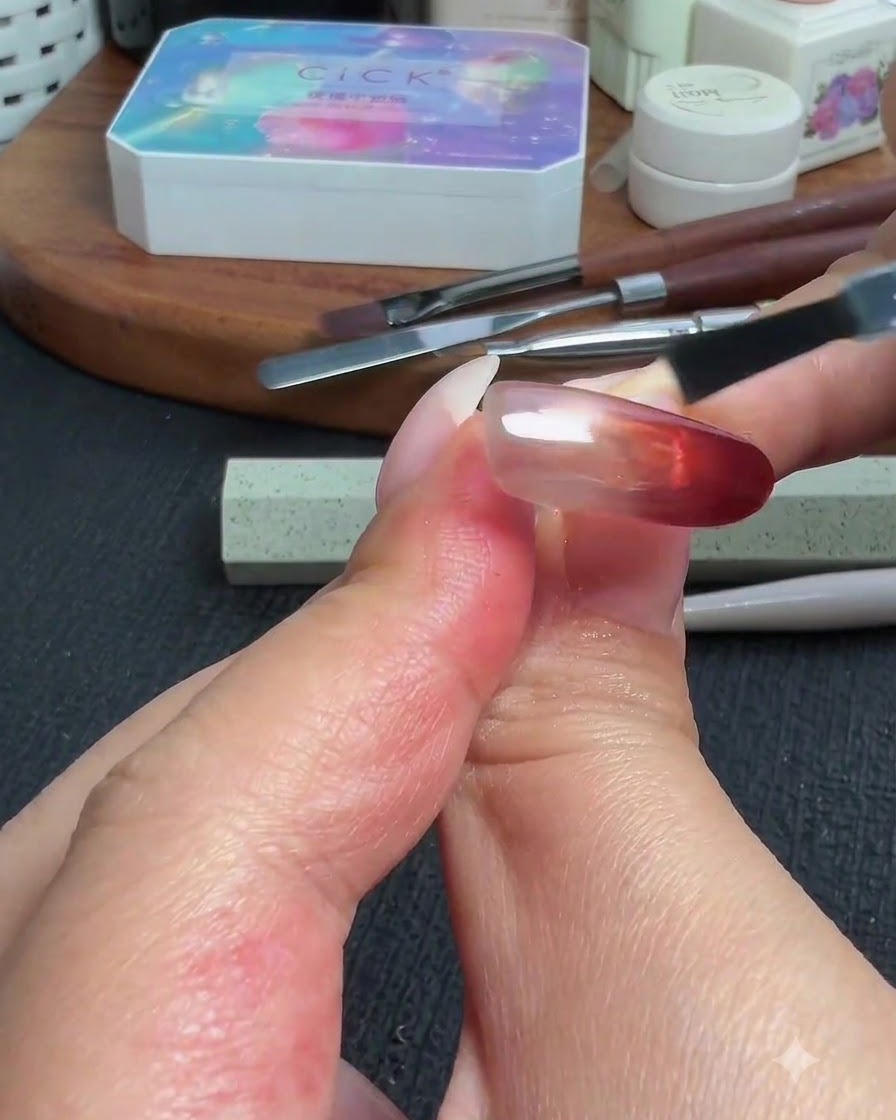 Un pinceau appliquant une couche de finition brillante sur longle terminé à dégradé rouge œil de chat et poudre.