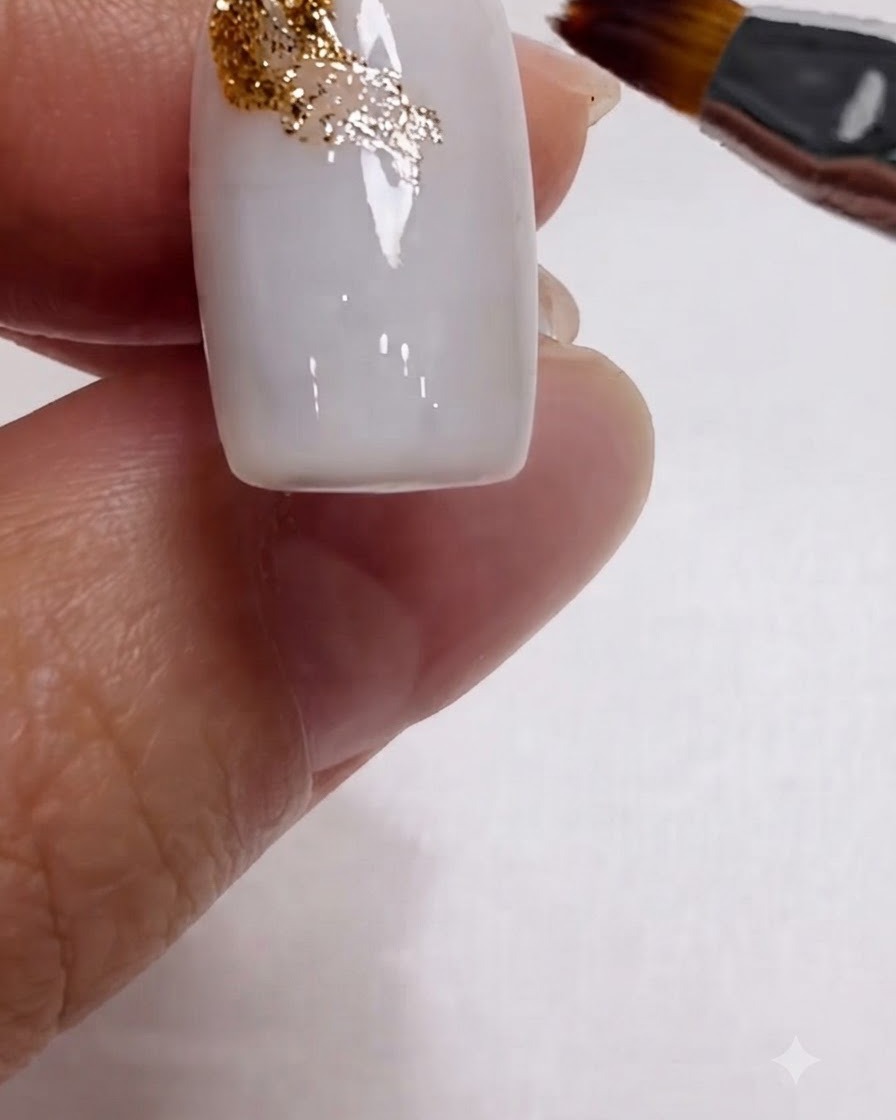Un pinceau estompant des paillettes dorées en diagonale sur le coin supérieur dun ongle blanc lait.