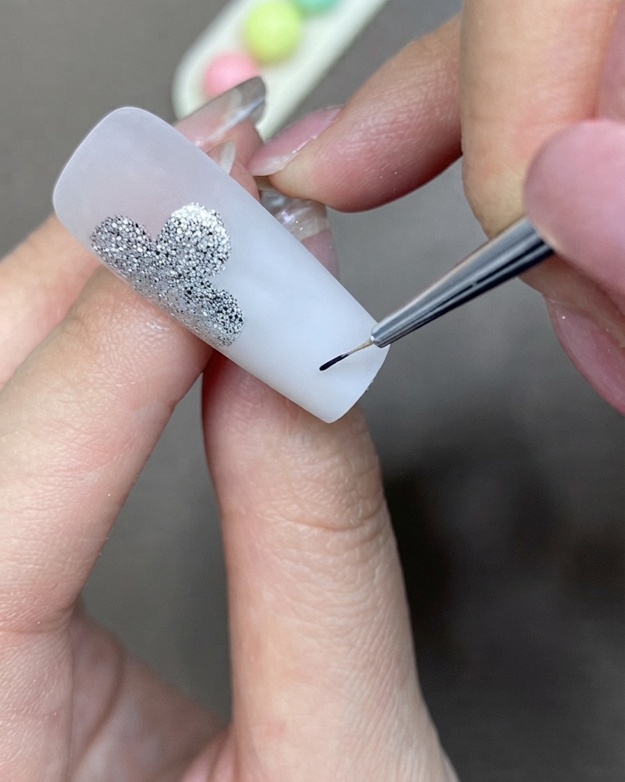 Un pinceau de détail fin appliquant du gel liner noir pour créer une forme de trèfle à côté dun design à paillettes argentées.