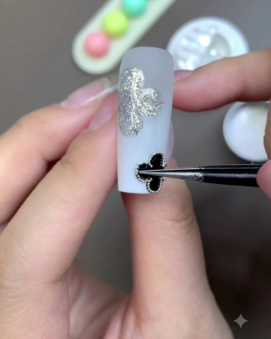 Une paire de pinces de précision plaquant un motif de trèfle noir encadré sur la surface dun ongle blanc laiteux.