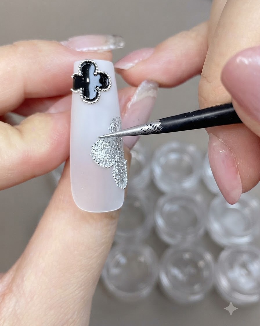 Un pinceau fin plaquant de petites perles dacier le long du bord dun design de trèfle pailleté argenté sur un ongle blanc.