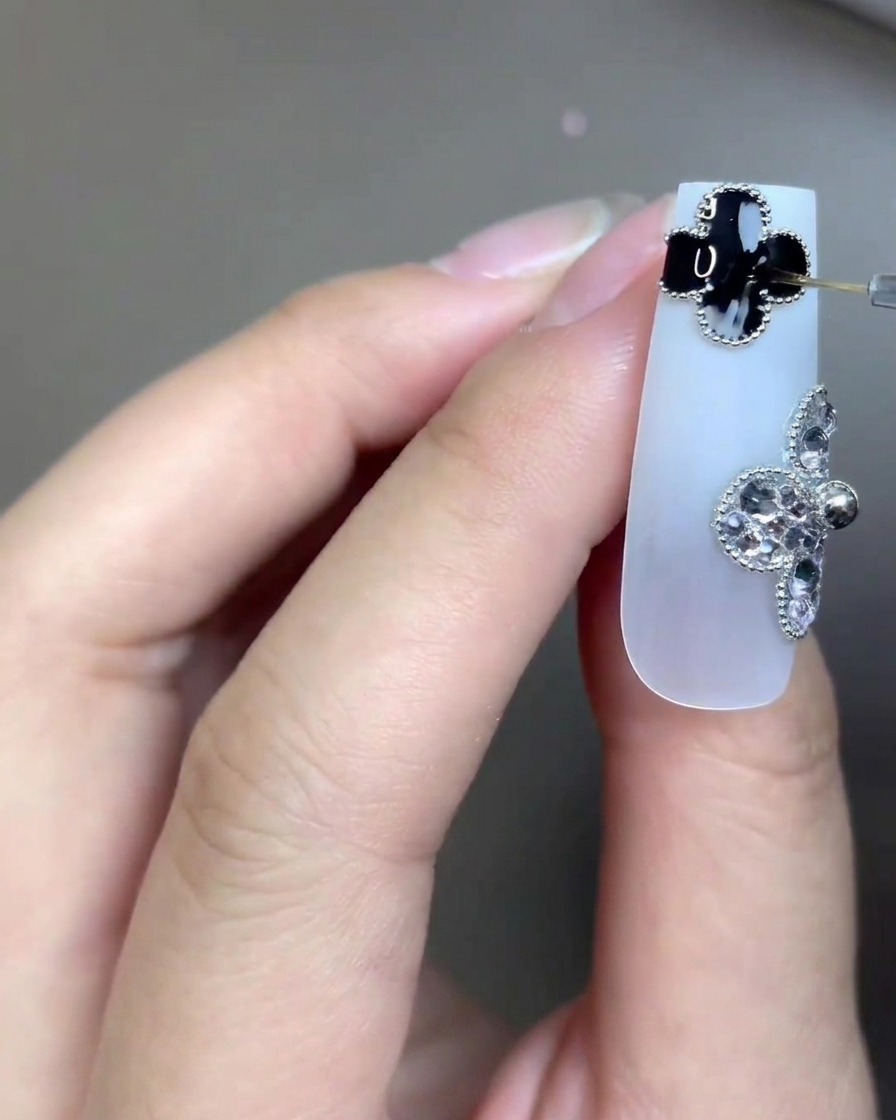 Gros plan dun ongle long blanc laiteux orné de deux charms en forme de trèfle à quatre feuilles — un noir, un en strass. Un pinceau très fin pour nail art applique un top coat transparent précisément sur le détail du trèfle noir.