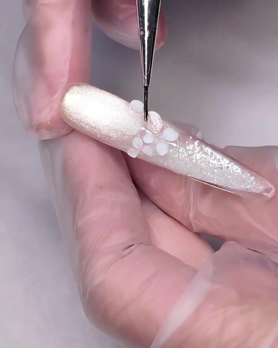 Un pinceau de détail fin appliquant du gel cat-eye rose nude sur une fleur sculptée en 3D blanche sur un ongle.