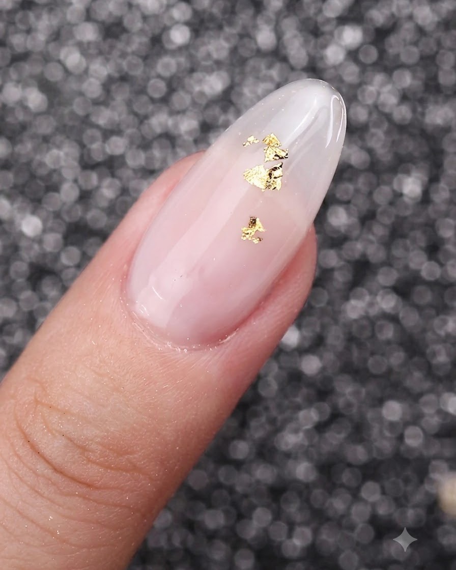 Une vue rapprochée de flocons de feuille dor délicatement posés sur un ongle manucuré au gel blanc laiteux.