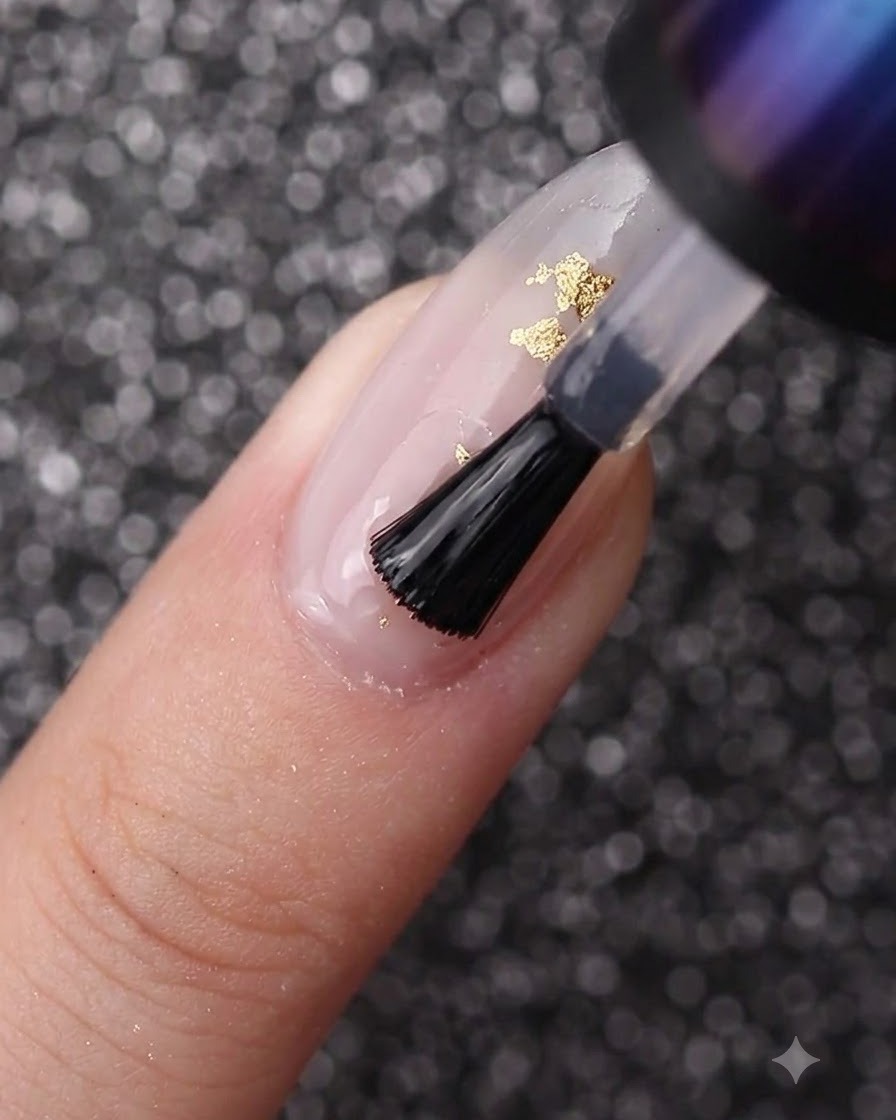 Un pinceau à ongles étalant du gel transparent sur un ongle décoré de vernis blanc laiteux et de feuille dor.