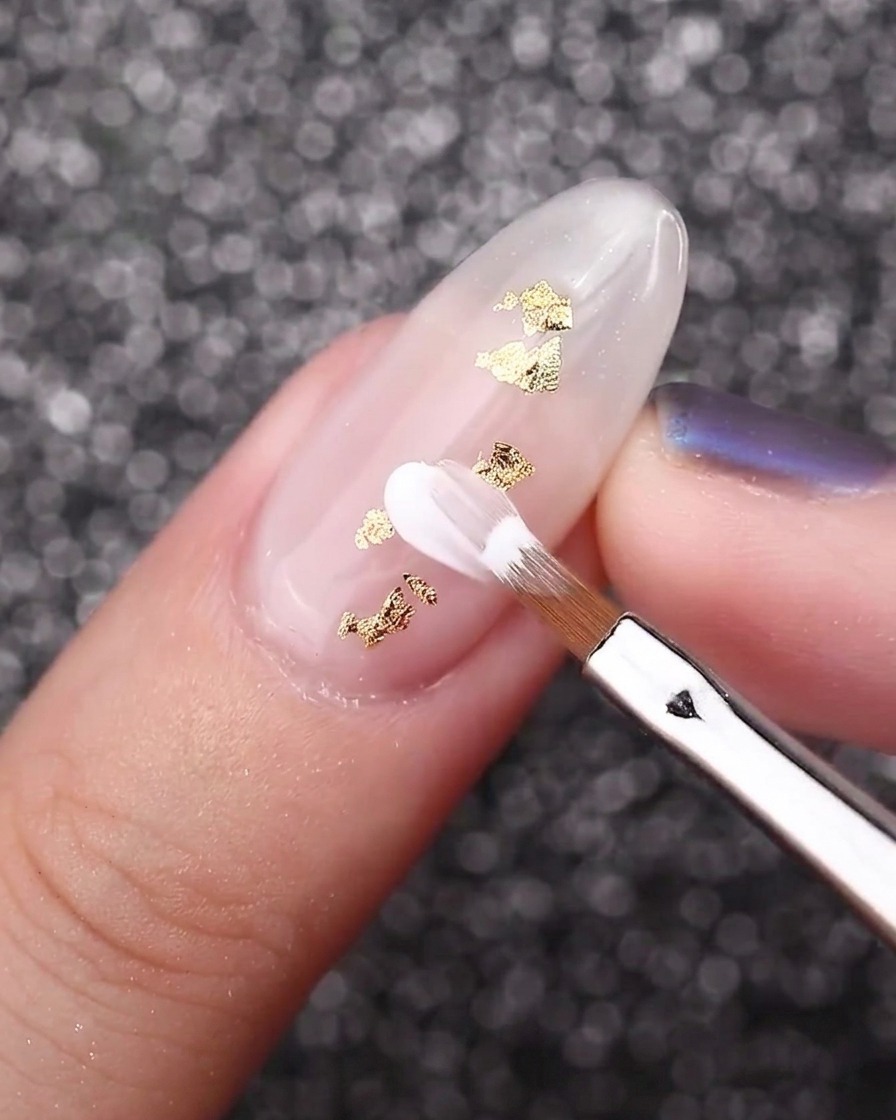 Un pinceau biseauté pour pétales créant des fleurs blanches sur un ongle blanc laiteux avec des accents de feuille dor.