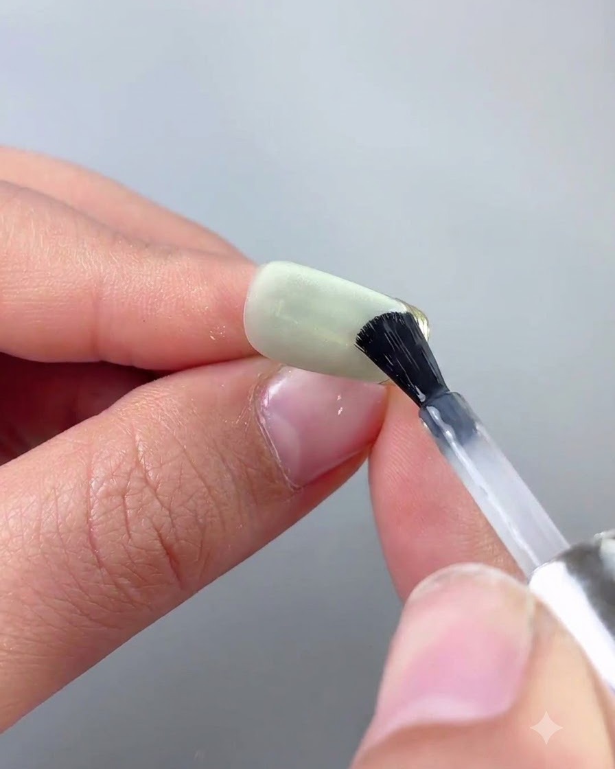 Un pinceau fin pour nail art appliquant précisément un top coat le long dun bord en chrome doré dun ongle cat eye vert.