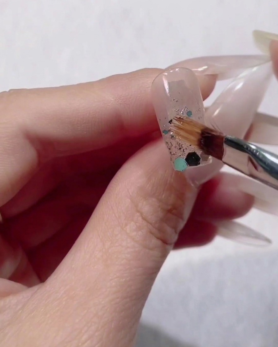 Un petit pinceau à ongle plaçant de grands sequins hexagonaux bleus et verts sur le dégradé de paillettes humides dun ongle.