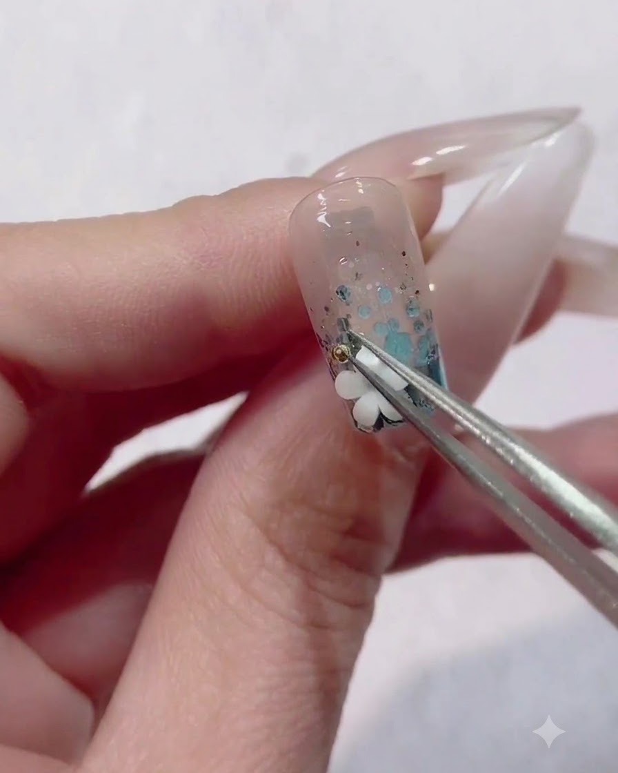 Une pince de précision plaçant une minuscule perle métallique à côté dun charm de fleur blanche sur un ongle décoré.