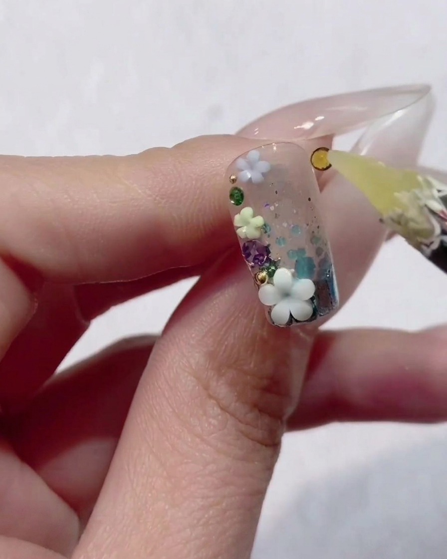 Gros plan dune main tenant un ongle décoré de gel transparent, de paillettes et de minuscules fleurs 3D, tandis quun outil à cire place un petit strass jaune dans le design.
