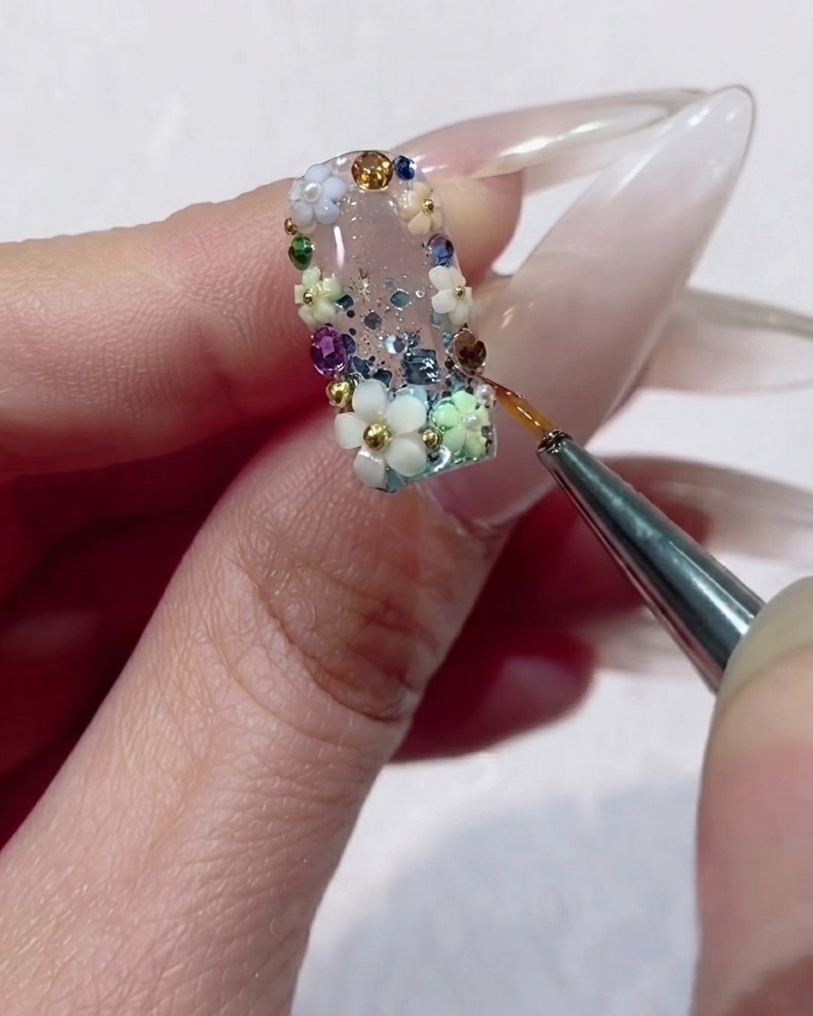 Un pinceau liner fin appliquant précisément du gel transparent autour des bords dun strass à facettes sur un design dongle floral.