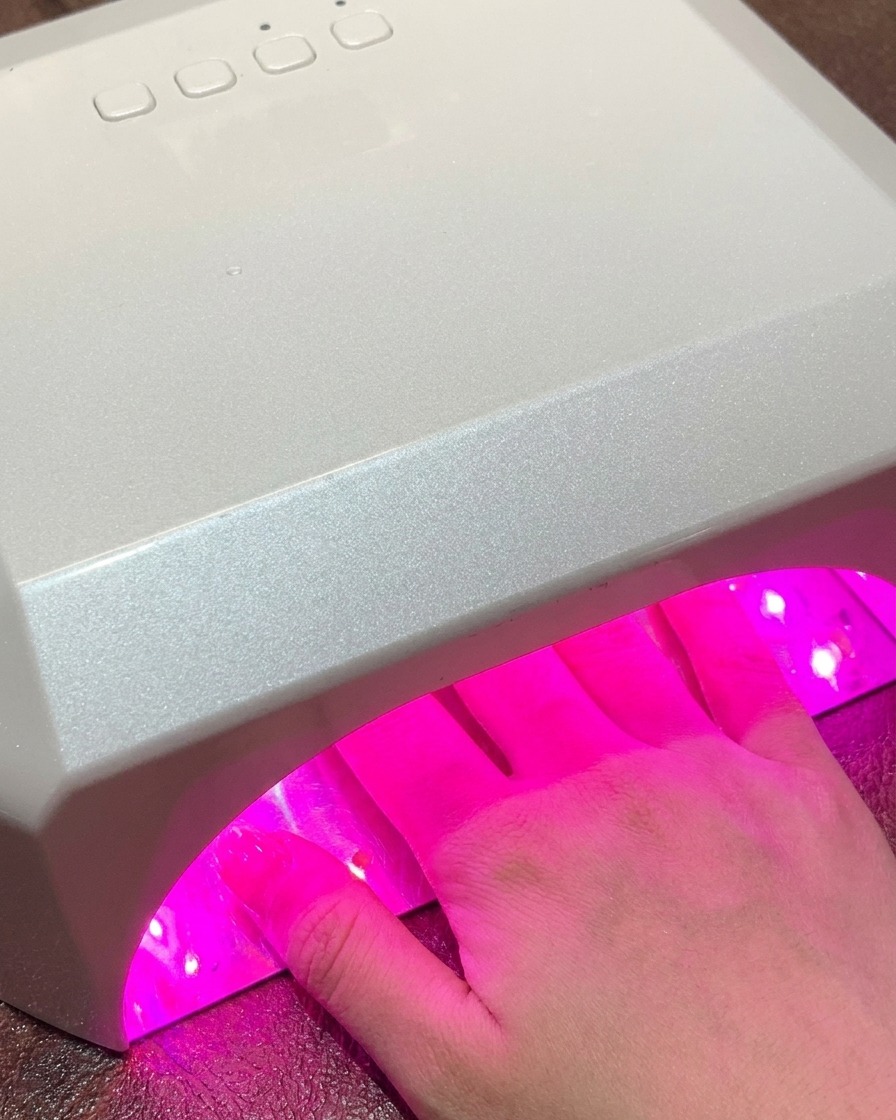 Une main placée à lintérieur dune grande lampe UV/LED standard pour ongles émettant une lumière rose pendant le processus de catalyse.