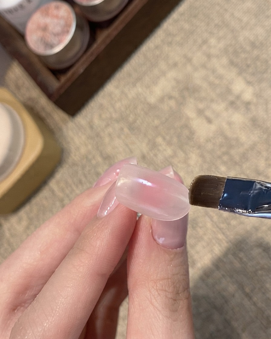 Un pinceau plat appliquant un gel cat-eye nacré bleu sur le bord dun ongle nude rosé.
