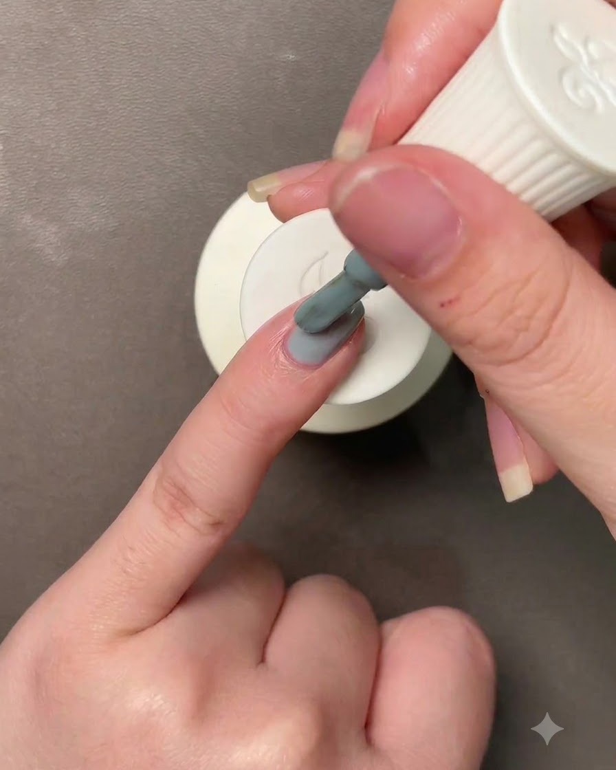 Une main tenant un pinceau appliquant un vernis gel gris bleu-vert Morandi sur un ongle.