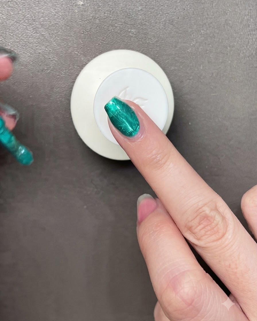 Un pinceau appliquant un vernis gel vert métallique vibrant sur un ongle avec un petit aimant visible à proximité.