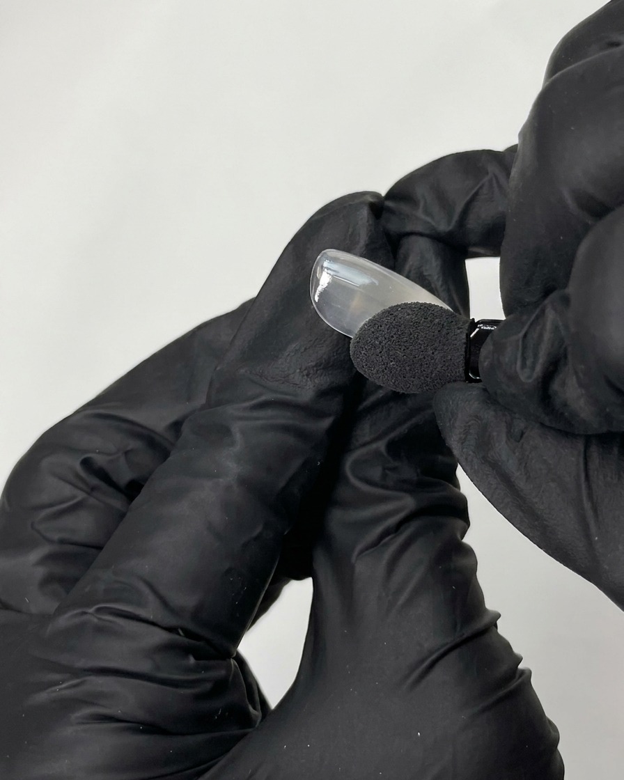 Une main gantée de noir utilisant un applicateur en mousse pour frotter de la poudre chromée irisée sur la pointe dun faux ongle lisse.