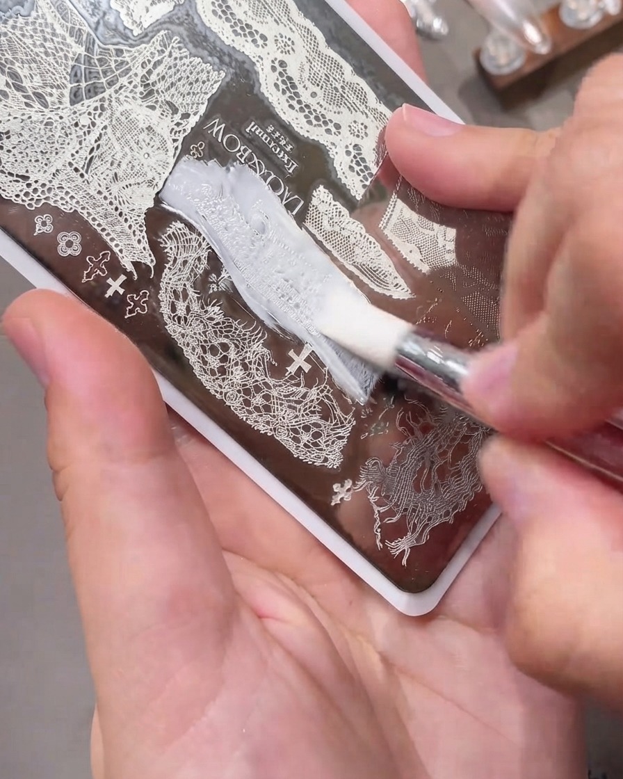 Une carte racloir glissant en douceur sur une plaque de stamping pour ongles en métal gravée remplie de vernis gel blanc.