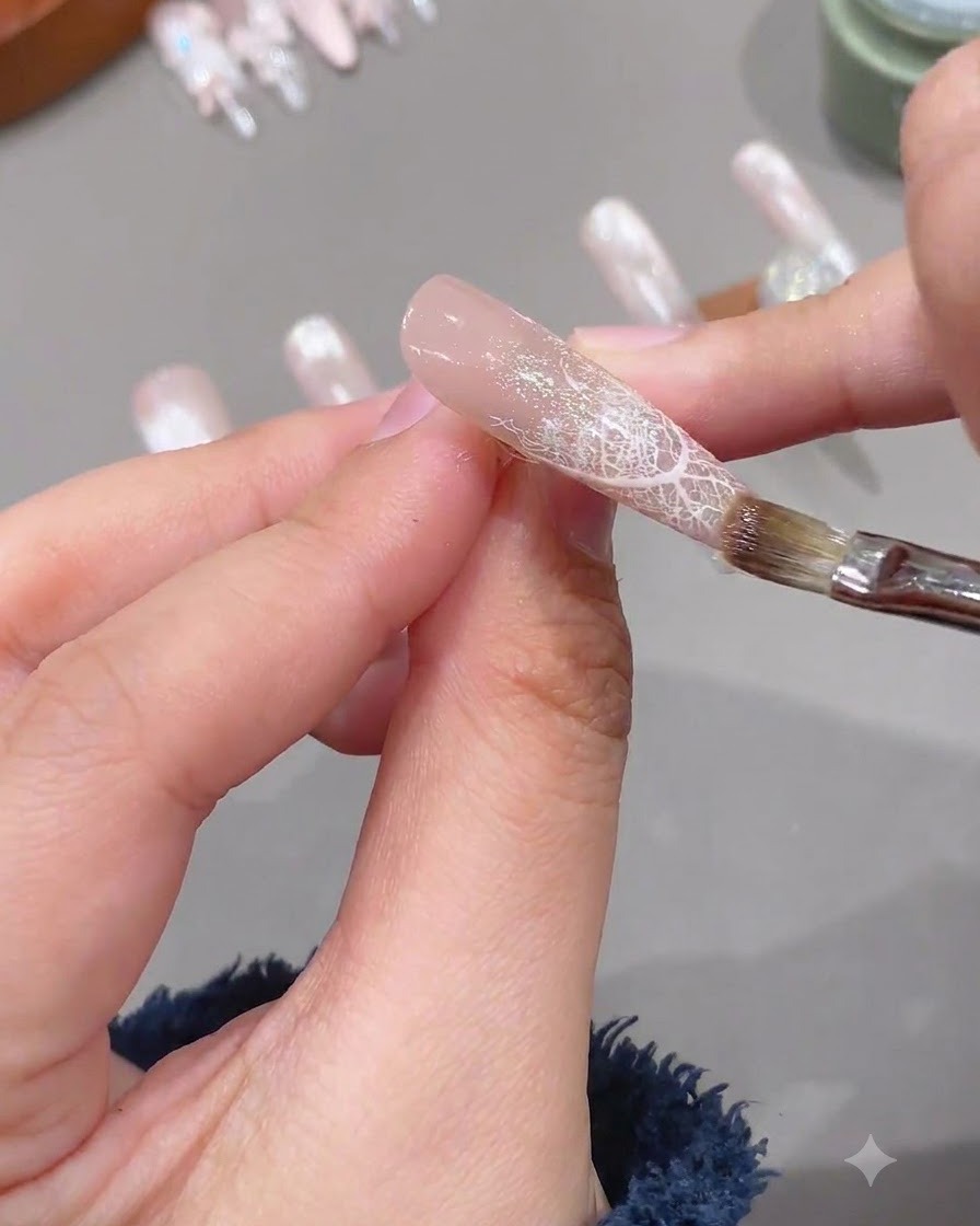 Un pinceau appliquant une couche transparente de gel de renforcement souple sur un ongle préparé avec un design en dentelle.