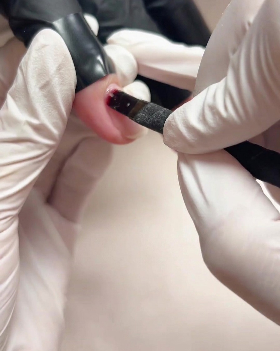 Gros plan dune main gantée utilisant un petit pinceau pour appliquer du gel de base transparent sur un ongle naturel.