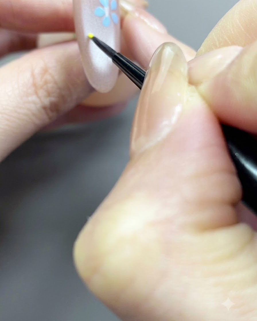 Un pinceau fin détaillant un petit point central jaune pour un design floral sur un ongle nude.