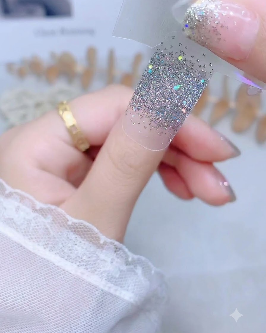 Une main tenant un autocollant dongle pailleté argenté par une languette de protection transparente au-dessus dun ongle de pouce nu.