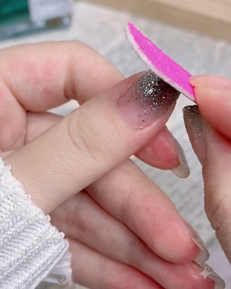 Une lime à ongles rose en carton limant la portion excédentaire dun sticker dongle pailleté argenté sur un pouce.