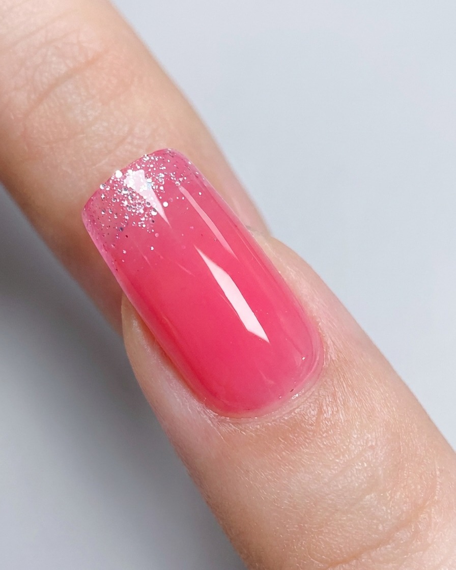 Gros plan sur un ongle rose brillant en dégradé avec un pot de gel pailleté argenté légèrement flou en arrière-plan.