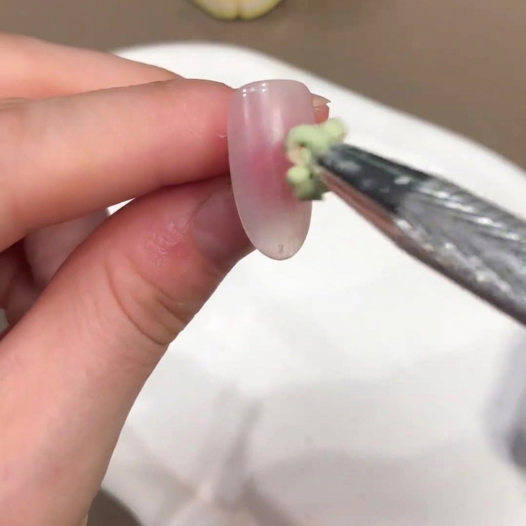 Un petit pinceau appliquant une goutte de gel teinté vert au centre dun ongle rose nude.
