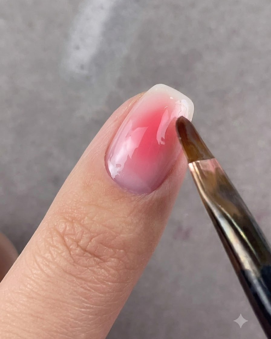 Un pinceau rond estompant délicatement les bords du gel rose sur un ongle.