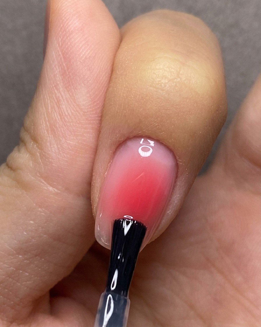 Un pinceau appliquant un top coat transparent uniformément sur un ongle avec un design en dégradé rose.