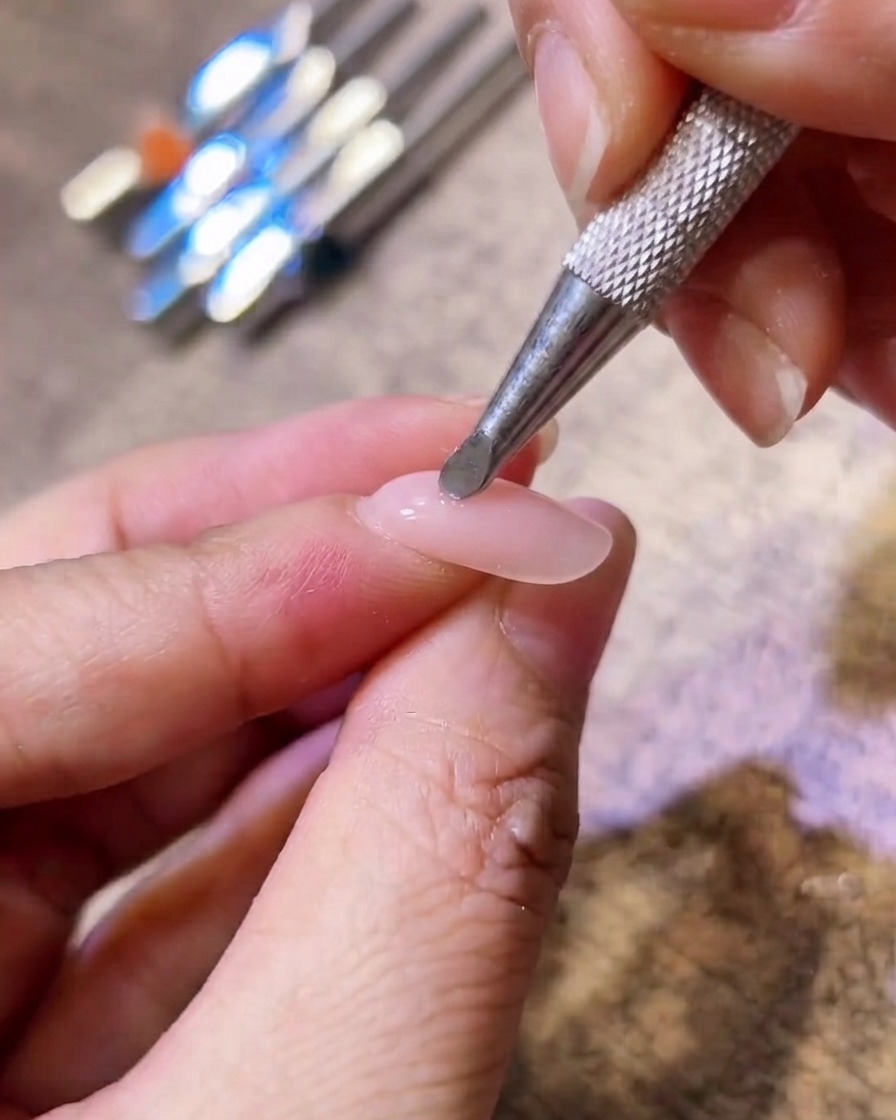 Gros plan dun outil applicateur en métal appliquant un pigment rose blush au centre dun ongle en gel nude.