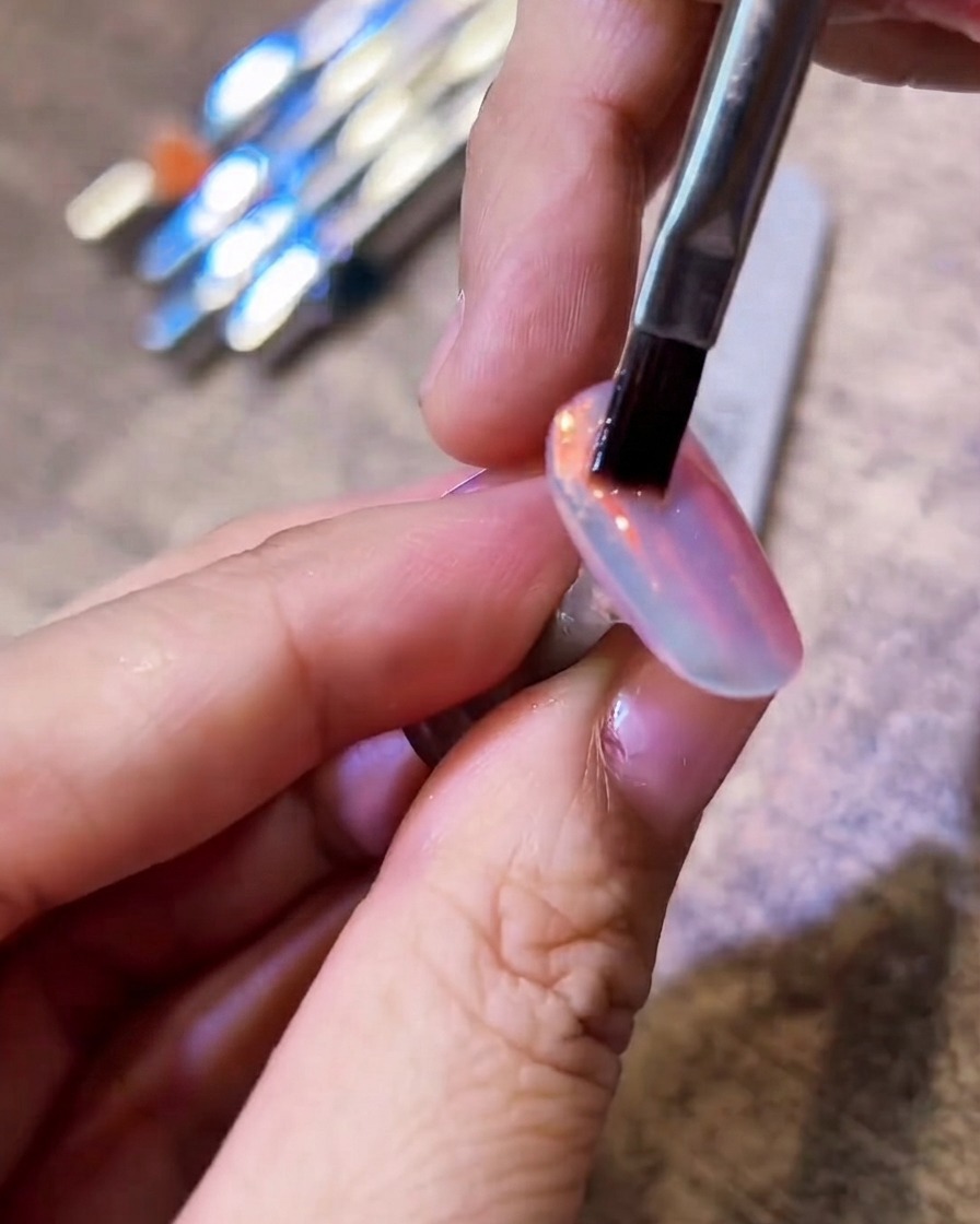 Un pinceau à poils plats balayant délicatement un ongle chrome aurora brillant pour éliminer lexcès de poudre.