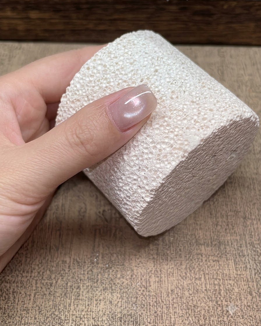 Gros plan sur un ongle unique posé contre un bloc blanc texturé, affichant un doux reflet cat eye perle de verre champagne.