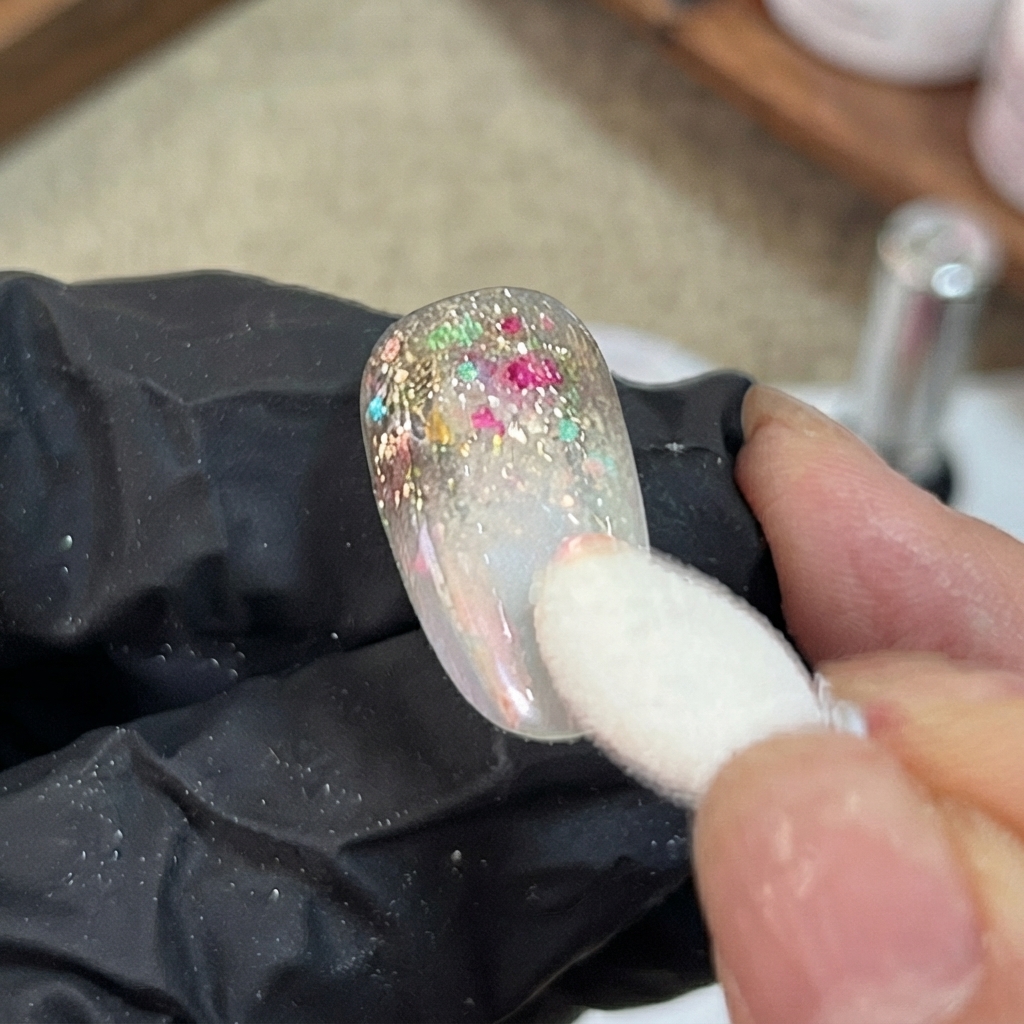 Un applicateur en éponge appliquant par tapotements de la poudre aurora iridescente sur la section dégradée dun faux ongle décoré.