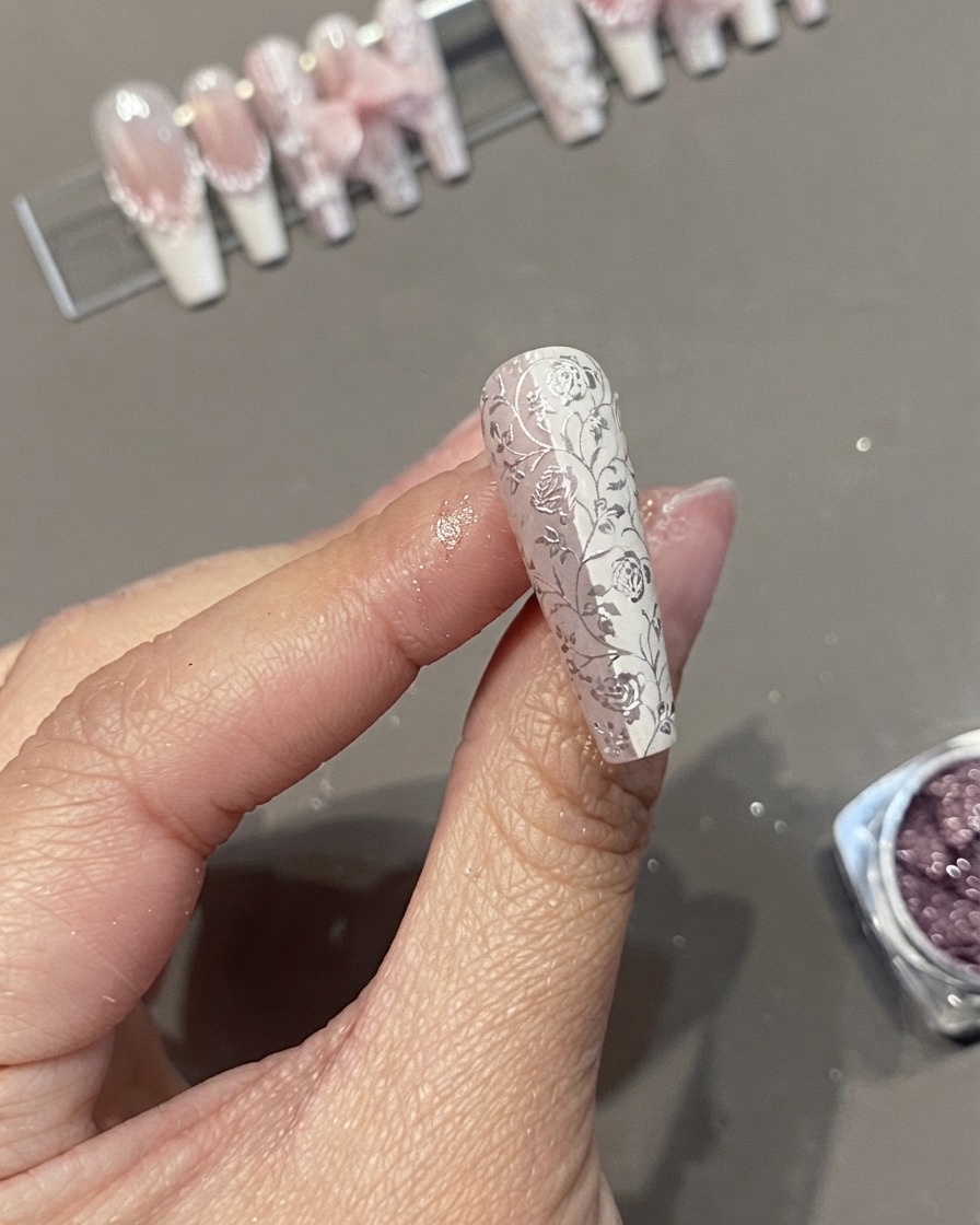 Gros plan dun faux ongle long en forme de cercueil orné dune dentelle florale blanche détaillée et dun motif chromé tenu entre les doigts.