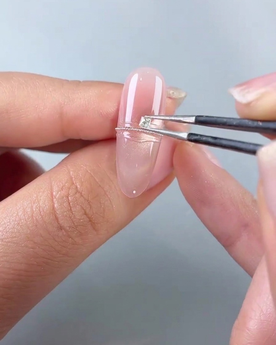 Une paire de brucelles plaçant un petit diamant carré à côté dune ligne de billes dacier sur un ongle en gel rose.