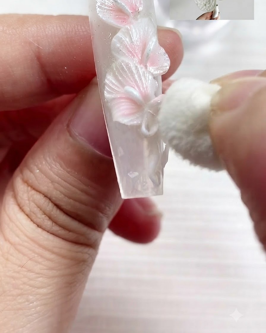 Une petite éponge cosmétique tamponnant délicatement du pigment blanc nacré sur les bords texturés de lys callas blancs sculptés et dun nœud sur un ongle.